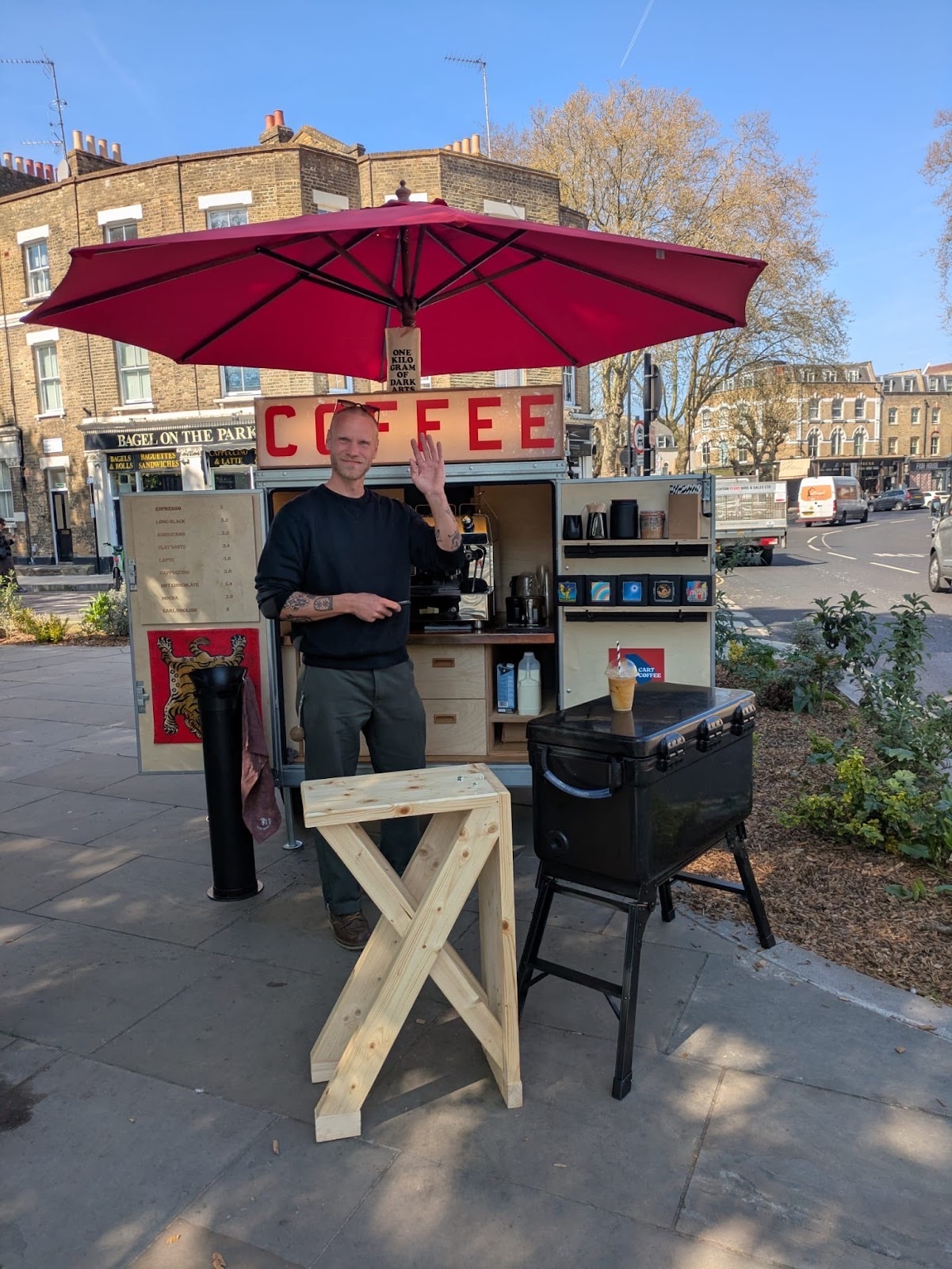 The open coffee cart with its espresso machine and wooden bar table under a red parasol on a sunny street